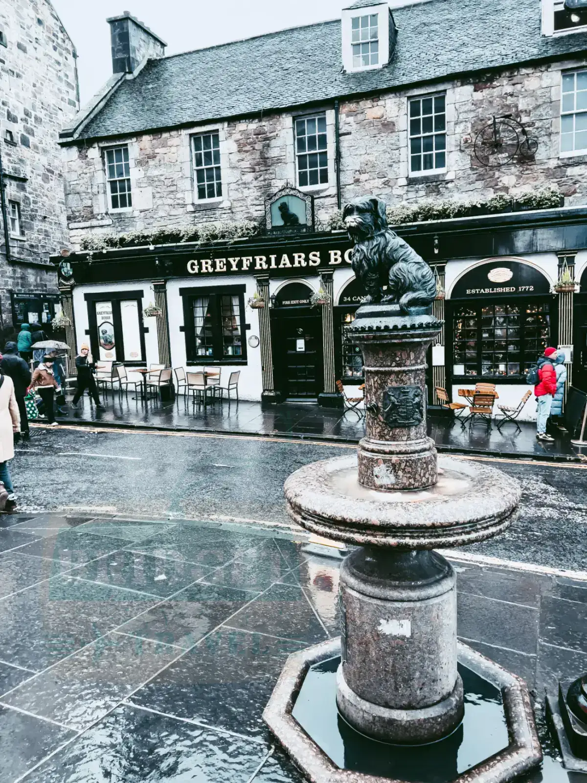 Greyfriars Bobby, Edynburg, Szkocja. Na pierwszym planie statuetka psa wraz z poidłem. Na drugim planie pub o nazwie Greyfriars Bobby.